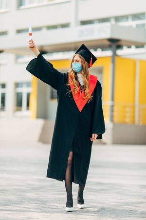 A female graduate student in a protective medical mask, in a black graduation dress, with a diploma in her hands. Graduation ceremony concept, quarantine, coronavirusの写真素材