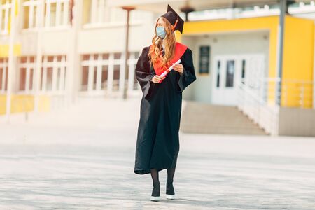 A female graduate student in a protective medical mask, in a black graduation dress, with a diploma in her hands. Graduation ceremony concept, quarantine, coronavirusの写真素材