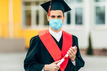 A male graduate student wears a protective mask against coronavirus, in a black graduation dress,...