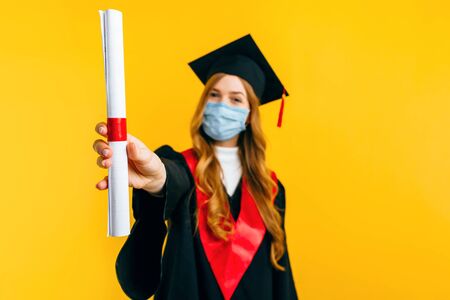 A happy, beautiful female graduate, wearing a medical protective mask, with a diploma on an isolated yellow background. Graduation, quarantine, coronavirusの写真素材