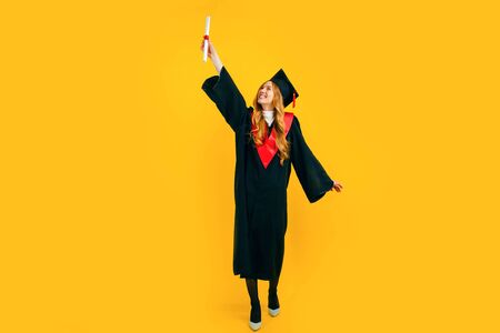 Full length, happy beautiful girl graduate happy and have fun with diploma on yellow background. The concept of the ceremonial presentation of diplomasの写真素材