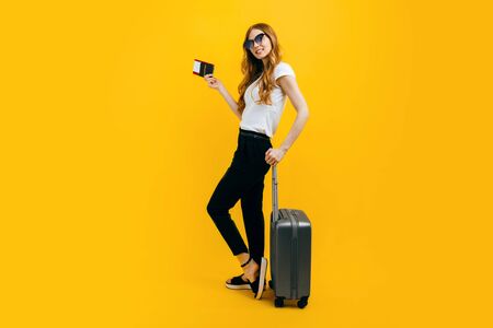 Happy beautiful woman, girl with a suitcase and passport with tickets on a yellow background. The concept of travel, businessの写真素材