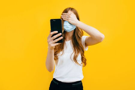 A young woman in a protective medical mask, using a smartphone and covering her eyes with her hands...