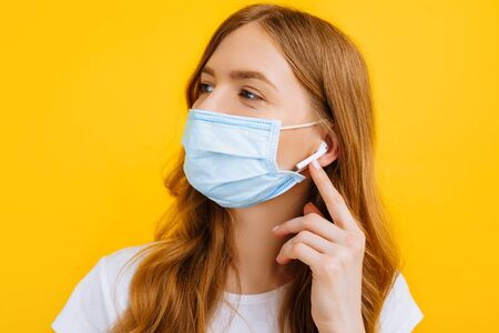 A young woman in a protective medical mask listens to music in wireless headphones on a yellow...
