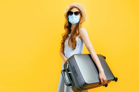 A young woman in a summer hat and a medical protective mask holds a suitcase in her hands on a blue background. The concept of quarantine, coronavirus, summer vacationの写真素材