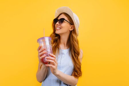 Happy beautiful girl in a hat and sunglasses with a refreshing summer cocktail on an isolated yellow background. Travel, vacation, summerの写真素材
