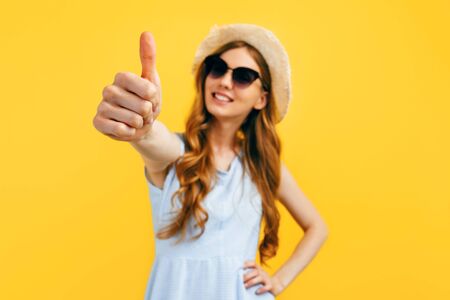 Cheerful beautiful girl in sunglasses and a summer hat, shows a gesture a thumb up, on a yellow background. Travel, vacation, summerの写真素材