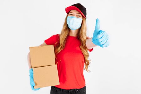 A uniformed courier, wearing medical rubber gloves and a medical mask, holds cardboard boxes and gives a thumbs-up gesture on a white background. Fast delivery. Online stores, payment by Bank transfer, quarantine, coronavirusの写真素材