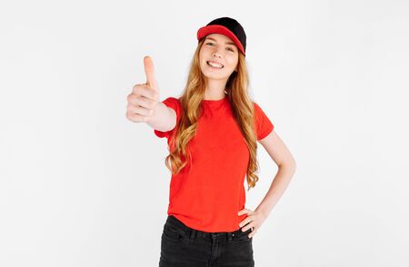 Happy courier girl in uniform, shows a thumbs up gesture on a white background. The concept of home deliveryの写真素材