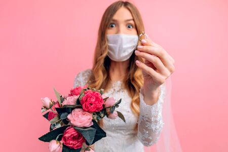 A bride in a wedding dress and a medical protective mask on her face shows her hand with an engagement ring on a pink background. Wedding, quarantine, coronavirusの写真素材