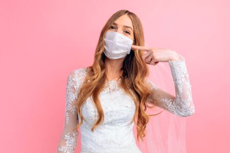 A bride in a wedding dress and a medical protective mask on her face points at the mask on a pink background. Wedding, quarantine, coronavirusの写真素材