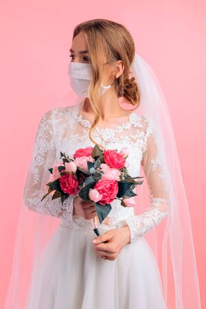 Portrait of a beautiful bride with a wedding bouquet in a medical protective mask on her face. Blonde with curly hair on a pink background. Quarantine, wedding, coronavirusの写真素材