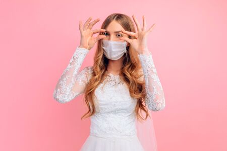 A beautiful bride in a wedding dress with a medical protective mask on her face, holding wedding rings on a pink background. Quarantine, wedding, coronavirusの写真素材