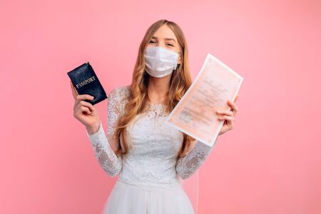 Beautiful bride in a wedding dress with a medical protective mask on her face, holding a passport and a marriage certificate, on a pink background. Quarantine, wedding, coronavirusの写真素材