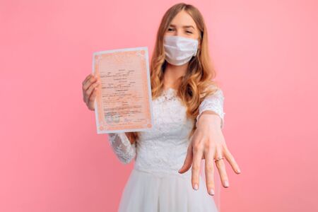 A beautiful bride in a wedding dress with a medical protective mask on her face, holding a marriage certificate, on a pink background. Quarantine, wedding, coronavirusの写真素材
