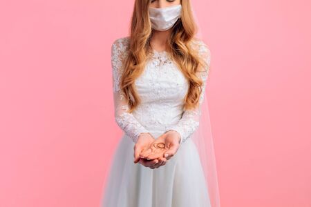 A beautiful bride in a wedding dress with a medical protective mask on her face, holding wedding rings on a pink background. Quarantine, wedding, coronavirusの写真素材