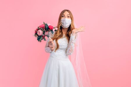 A happy bride with a bouquet of flowers in a wedding dress and a medical protective mask on her face, on a pink background. Wedding, quarantine, coronavirusの写真素材