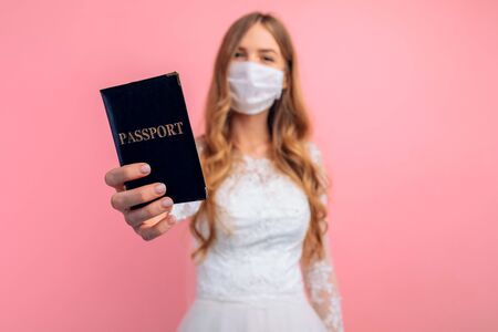 A beautiful bride in a wedding dress with a medical protective mask on her face, holding a passport in her hand, a blonde with curly hair on a pink background. Quarantine, wedding, coronavirusの写真素材