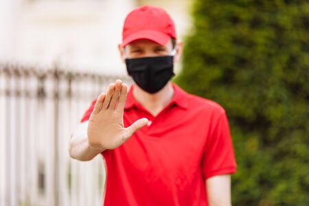 A courier in uniform and wearing a medical protective mask, shows a stop gesture. Fast delivery by transport. Online stores and Express delivery. Quarantine, coronavirusの写真素材