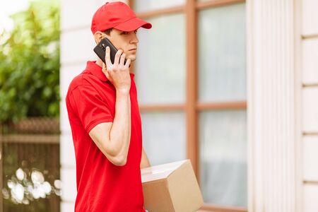 Young happy courier with a cardboard box, talking on a mobile phone. Home deliveryの写真素材