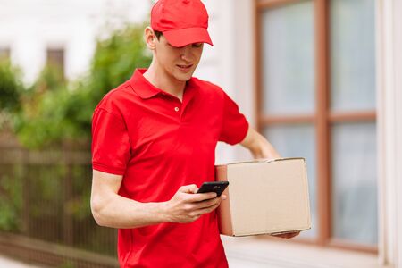 A young happy courier with a cardboard box, using a mobile phone. Home deliveryの写真素材