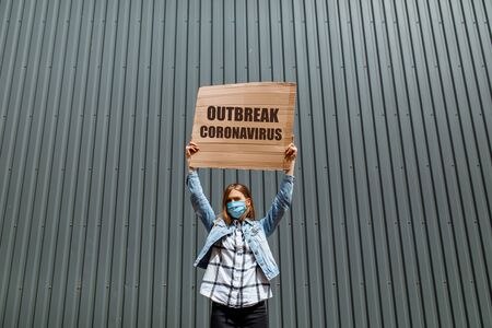 A young woman in a medical protective mask holds a cardboard poster with the words "coronavirus Outbreak", standing in the city against a gray wall. Quarantine, coronavirusの写真素材