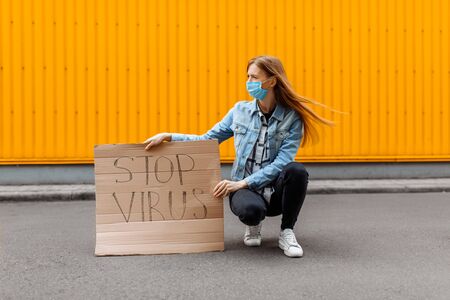 A young woman in a medical protective mask holds a cardboard placard with the words STOP the VIRUS, sitting against a yellow wallの写真素材