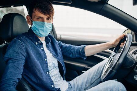 A young man in a protective medical mask, sitting at the wheel of a car, the driver is driving. Quarantine, coronavirusの写真素材