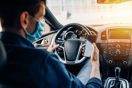 A man in a protective medical mask, sitting behind the wheel of a car, uses a mobile phone while...
