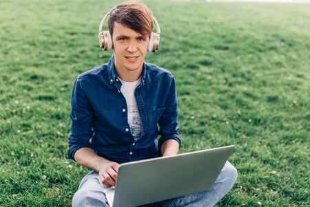 A young handsome man is sitting outside on the grass with a laptop and listening to music with headphonesの写真素材