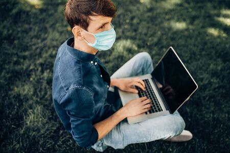 A young student in a protective medical mask sits on the grass in a Park with a laptop. Quarantine, coronavirusの写真素材