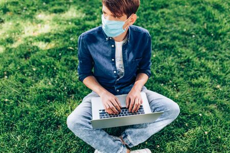 A young student in a protective medical mask sits on the grass in a Park with a laptop. Quarantine, coronavirusの写真素材
