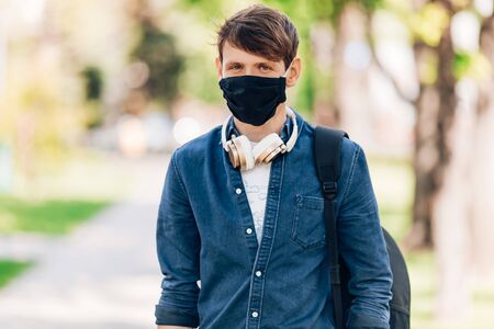 A young man in a protective medical mask on his face walks through the Park with wireless headphones...