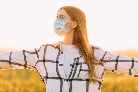 beautiful woman in a checked shirt and a protective medical mask on her face stands in a wheat field at sunset. Quarantine, coronavirus, cropの写真素材