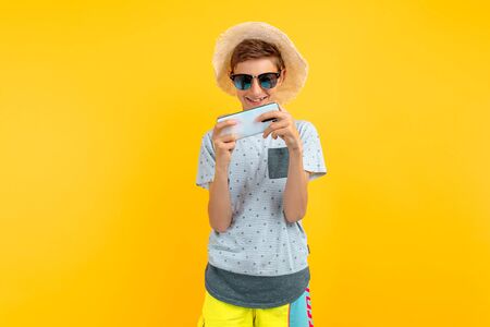 Stylish smiling teen guy in a summer hat using a mobile phone standing on an isolated yellow...