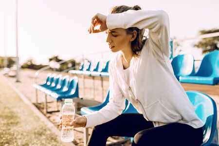 A tired sportswoman relaxes after training, holding a bottle of water in her hands, a woman rests...