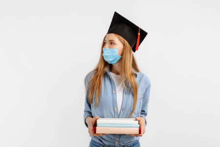 graduate girl in a medical mask on her face and a graduation hat on her head, with books in her hands, stands on a white background. Graduation distance educationの写真素材