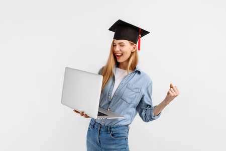 Happy excited graduate girl in a graduation cap on her head using a laptop and showing a winning gesture while standing on a white background. Graduation, distance learningの写真素材