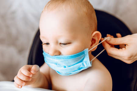 mother puts a medical protective mask on the face of her young child, the Child is sitting in a feeding chair. Quarantineの写真素材