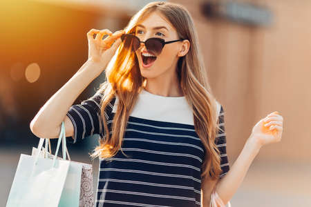 Excited shaken happy stylish young woman in sunglasses, with colorful shopping bags, shopping, standing on the street in the background of the shopping center. Shopping, fashionの写真素材