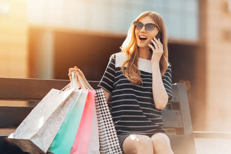 happy beautiful young woman wearing sunglasses is sitting on a bench in the background of a shopping center with shopping bags, talking on a mobile phone. Shopping, fashionの写真素材