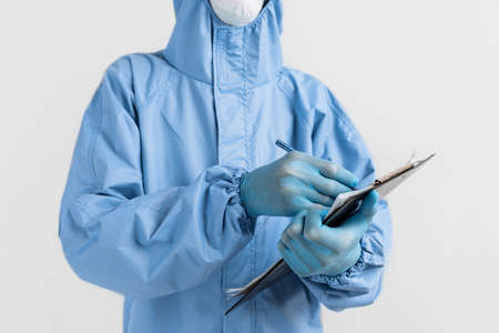 doctor, dressed in a protective suit against coronavirus, glasses, mask and gloves, writes documents...