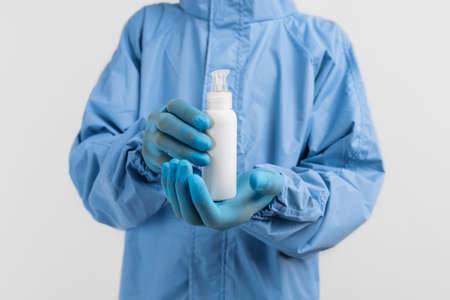 doctor in a protective medical suit against coronavirus, wearing latex gloves, holds a jar of antiseptic disinfectant on an isolated white background. Coronavirus, quarantineの写真素材