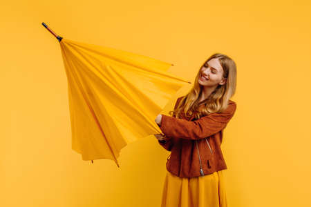 happy attractive girl in a yellow dress and autumn jacket opens a yellow umbrella on an isolated yellow backgroundの写真素材