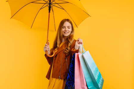 happy girl, in a bright yellow dress and autumn jacket, standing with a yellow umbrella and shopping bags, on an yellow backgroundの写真素材