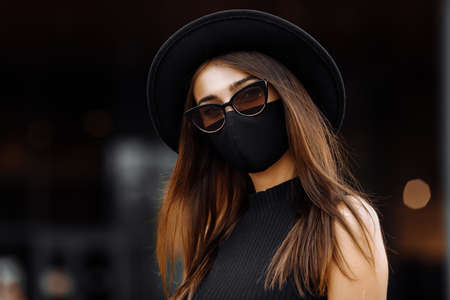 beautiful business woman in a black medical protective mask on her face, dressed in a black elegant dress and hat, wearing dark glasses, poses standing against the background of a glass building. Businessの写真素材