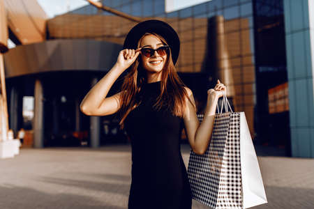 Happy attractive elegant girl, in a black dress and hat, wearing dark glasses, holding shopping bags and enjoying shopping. Black Fridayの写真素材