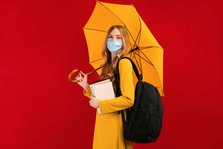 female student in a medical protective mask on her face, with a backpack and books, and a yellow...