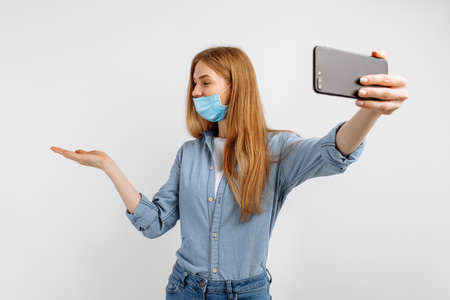 young woman in a medical protective mask, talking on a mobile phone video link, on a white backgroundの写真素材