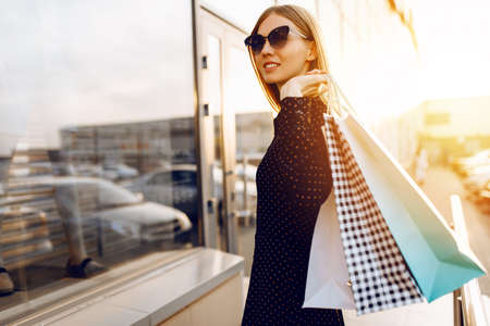 young woman in sunglasses and dress, with shopping bags, in the city against the background of a shopping center at sunsetの写真素材